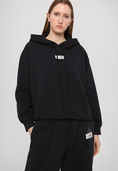 Svart kort hoodie med dragsko, ficka fram och en etikett på bröstet. Mjukt tyg med avslappnad passform och ribbade ärmslut.