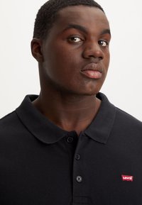 Tamsiai mėlynas polo marškinėlis, pagamintas iš struktūruotos medžiagos. Turi apykaklę, du mygtukus ir raudoną Levi's logotipą kairėje krūtinės pusėje.