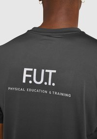 Svart atletisk skjorta med vit text "F.U.T." och "FYSISK UTRUSTNING & TRÄNING" på baksidan. Slät textur, korta ärmar.