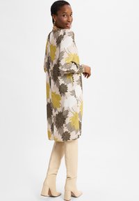 Frau in einem beigen, floral gemusterten Langarmkleid mit gelben und grauen Blumen, kombiniert mit cremefarbenen kniehohen Stiefeletten mit Absatz, die über die Schulter blickt.