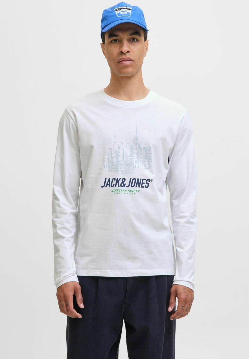 Man in een wit overhemd met lange mouwen met een stadsgezicht en de tekst "JACK & JONES", een blauwe pet en donkere broek tegen een effen achtergrond.