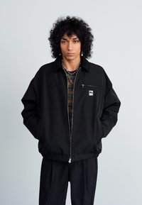 Jovem com cabelo encaracolado, vestindo uma jaqueta preta com fecho éclair sobre uma camisa às riscas, de pé com as mãos nos bolsos, contra um fundo simples.