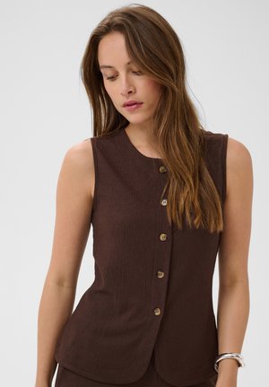 Femme aux longs cheveux bruns portant un top marron sans manches boutonné et un bracelet argenté à son poignet gauche, regardant vers le bas.