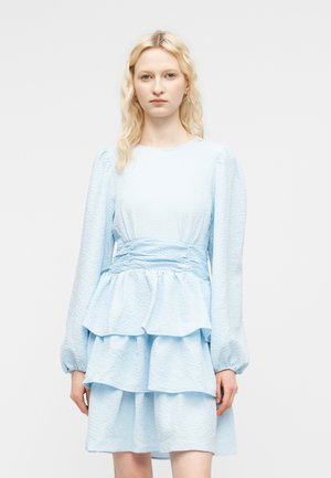 Lyshåret kvinde iført en lyseblå, tekstureret, lagdelt kjole med lange ærmer og rynket talje, stående foran en ensfarvet hvid baggrund.