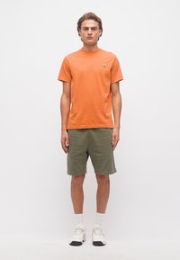 Νεαρός άνδρας που φοράει πορτοκαλί κοντομάνικο μπλουζάκι, χακί σορτς, λευκές κάλτσες και λευκά αθλητικά παπούτσια, στέκεται μπροστά σε απλό λευκό φόντο.