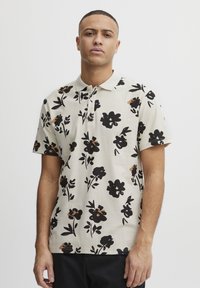Pikéskjorte med korte ermer i beige stoff med svarte blomstermønstre og brune detaljer, samt krage.