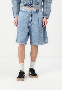 Bermudas de mezclilla azul claro con un ajuste relajado, con pliegues en la cintura y bolsillos laterales. Combinadas con zapatillas negras y calcetines blancos de caña baja.
