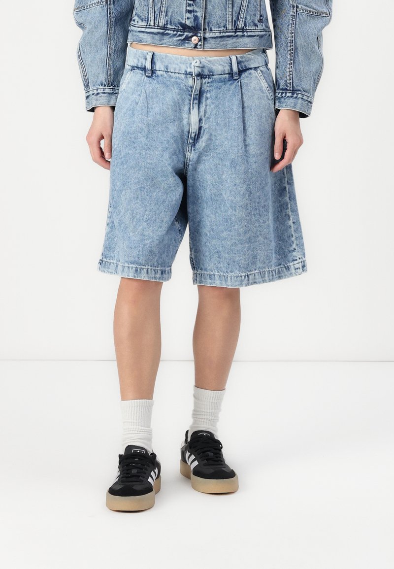 Bermudas de mezclilla azul claro con un ajuste relajado, con pliegues en la cintura y bolsillos laterales. Combinadas con zapatillas negras y calcetines blancos de caña baja.