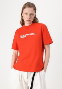 Rdeča majica s kratkimi rokavi iz bombaža, ki ima bel napis "KARL LAGERFELD JEANS" čez prsni del, okroglo ovratnik ter sproščen kroj.
