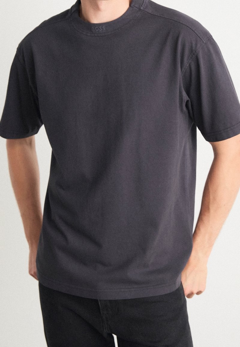 Homme portant un t-shirt gris foncé uni à manches courtes avec un petit logo en relief "BOSS" sur le col et un pantalon noir.