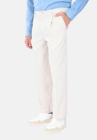 Pantaloni plissettati color beige chiaro, realizzati in un tessuto liscio. Presentano tasche anteriori e una chiusura con un singolo bottone. Indossati con sneaker bianche.