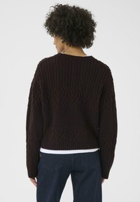 Tmavě hnědý pletený svetr s texturovaným vzorem, s kulatým výstřihem a oversized rukávy. Nosený nad tmavými džínami.