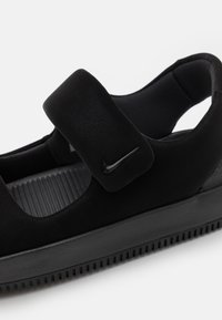 Zapatilla negra con parte superior texturizada, cinta de velcro y suela de goma con dibujo. Presenta un logo sutil en la cinta como marca.