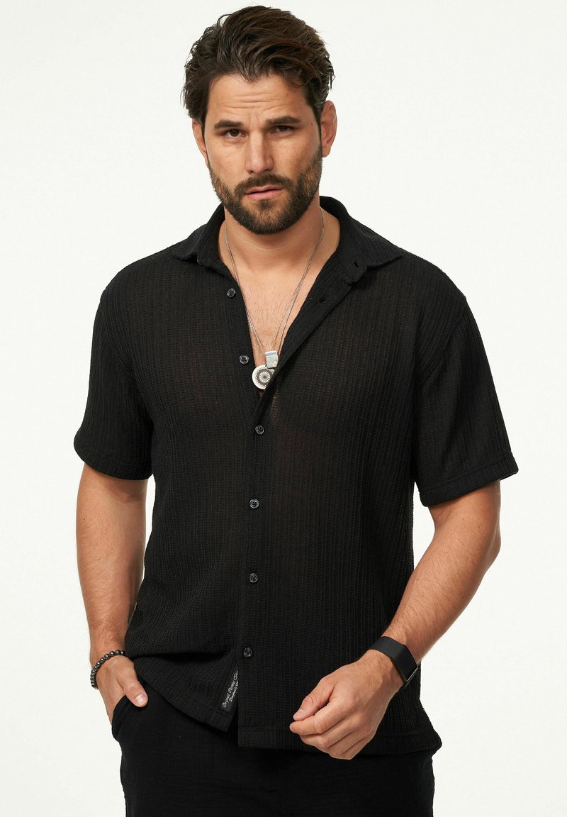 Schwarzes kurzärmeliges Hemd aus strukturiertem, geripptem Stoff. Verfügt über eine durchgehende Knopfleiste und einen lässigen Kragen. Wird mit einer Anhänger-Halskette getragen.