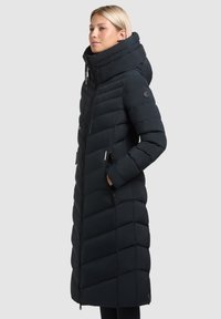 Abrigo puffer largo negro con un cuello alto, diseño acolchado horizontal, cierre de cremallera y bolsillos laterales. La tela parece suave y texturada.