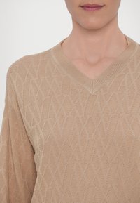 Maglione beige lavorato a maglia con scollo a V, caratterizzato da motivi geometrici, finitura testurizzata e maniche lunghe. Il tessuto sembra leggero e traspirante.