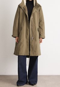 Parka verde oliva con cappuccio foderato in pelliccia sintetica, chiusure a scatto e zip, tasche laterali e una silhouette rilassata, abbinata a jeans a gamba larga blu scuro.