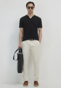 Camicia nera a maniche corte, pantaloni crema, scarpe in suede marroni, tiene una borsa a tracolla nera, in piedi su uno sfondo bianco semplice.