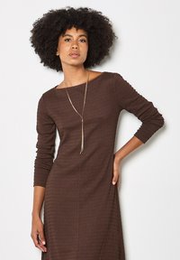 Brown ribbed dress with three-quarter sleeves and a boat neckline. Features a gold pendant necklace and textured fabric.