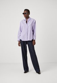 Lavender striped button-up shirt with long sleeves, semi-relaxed fit, paired with dark tailored pants and black sunglasses.