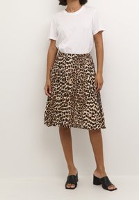 Kobieta w białej, krótko rękawowej koszulce, w spódnicy plisowanej w panterkę oraz czarnych, otwartych na palce klapach na obcasie stoi pod jednolitą ścianą.