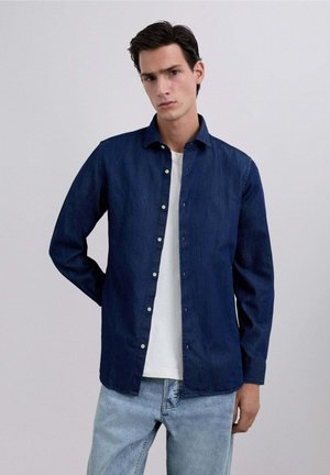 Camisa de mezclilla azul oscuro con diseño de botones, de manga larga y cuello clásico, llevada sobre una camiseta blanca, combinada con jeans claros.