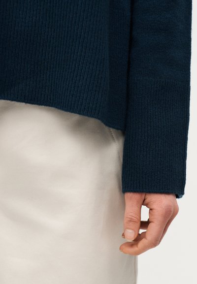 Χέρι που φοράει μανίκι σκουρόχρωμου μπλε πλεκτού πουλόβερ, ξεκουράζεται δίπλα σε μπεζ παντελόνι από ύφασμα.