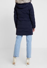Giacca a vento blu navy con cappuccio foderato in pelliccia, caratterizzata da un design trapuntato e maniche lunghe. Abbinata a jeans in denim azzurro chiaro e tacchi beige.