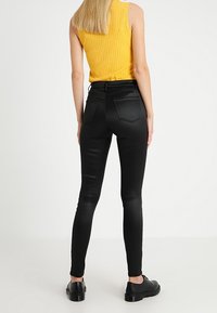 Černé slim-fit džíny s hladkým povrchem, obsahující dvě zadní kapsy. Model má na sobě žlutý hladký tílko a černé boty.