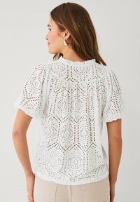 Hvid bomuldsbluse med puffærmer, der har intrikate hulbroderier og bølgende kanter. Rynket halsudskæring med en løs pasform.