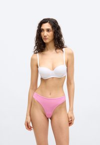 Γυναίκα με σγουρά σκουρόχρωμα μαλλιά που φοράει λευκό σουτιέν χωρίς τιράντες και ροζ εσώρουχο με ψηλή γραμμή σε απλό λευκό φόντο.