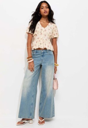 Vrouw draagt een cremekleurige blouse met bloemenpatroon, wijde lichtblauwe spijkerbroek, witte sandalen met hak en meerdere armbanden, en houdt een kleine roze handtas vast.