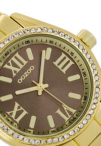 Montre-bracelet en métal doré avec un cadran marron, des index horaires en chiffres romains et des accents en cristal sur la lunette. Elle est dotée d'un bracelet en acier inoxydable.