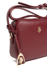 Bordowa torebka na ramię ze skóry o fakturowanej powierzchni, złotymi elementami i zawieszką z logo. Posiada regulowany pasek i zamknięcie na zamek.