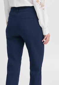 Marineblaue maßgeschneiderte Hose mit einer enganliegenden Form, Rückentaschen-Detaillierung und einem glatten, leicht strukturierten Stoff. Das Modell trägt eine weiße Bluse.