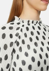 Witte transparante blouse met grote zwarte stippen. Heeft een gerimpelde halslijn en verticale plooien, wat een zachte, lichte textuur benadrukt.