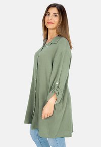 Grünes, strukturiertes Hemd mit Kragen, aufgerollten Ärmeln und lockerer Passform. Kombiniert mit hellblauen Jeans.