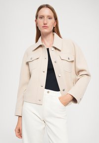 Giacca beige realizzata in morbido tessuto, con colletto, chiusura a bottoni, due tasche sul petto e un design cropped. Abbinata a pantaloni bianchi.