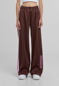 Pantalones de chándal anchos marrones con cinturilla elástica, que presentan rayas laterales rosas y blancas y el texto "FELICIOUS" en la parte inferior de la pierna izquierda.
