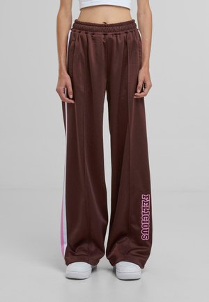 Bruine brede sweatpants met een elastische tailleband, voorzien van roze en witte zijstrepen en de tekst "FELICIOUS" op de onderkant van het linkerpijpje.