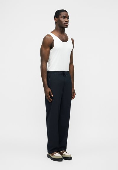 Αθλητικός άνδρας όρθιος, φορώντας λευκό αμάνικο μπλουζάκι, μαύρο παντελόνι με κορδόνια και μπεζ και καφέ casual παπούτσια, μπροστά σε απλό λευκό φόντο.