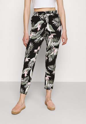 Femme portant un pantalon noir fuselé avec des motifs de feuilles tropicales vertes et roses, assorti à des chaussures slip-on roses pâles et un crop top blanc.