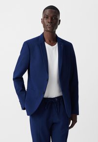 Granatowa marynarka z teksturowanej tkaniny, z pojedynczym guzikiem, klapami w kształcie notcha i bocznymi kieszeniami, noszona na białej koszuli.
