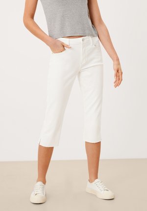Femme portant un pantalon blanc court avec des ourlets bruts, un haut gris sans manches et des baskets blanches, debout avec une main dans la poche.