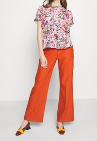 Blusa floral de manga corta en tonos rosa, verde y púrpura, combinada con pantalones anchos naranjas y zapatos de tacón bajo multicolores.