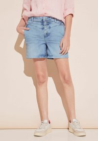 Pantalones cortos de mezclilla azul claro con cierre de cinco botones, bolsillos laterales y un ajuste casual, combinados con zapatillas blancas con acentos rosas.
