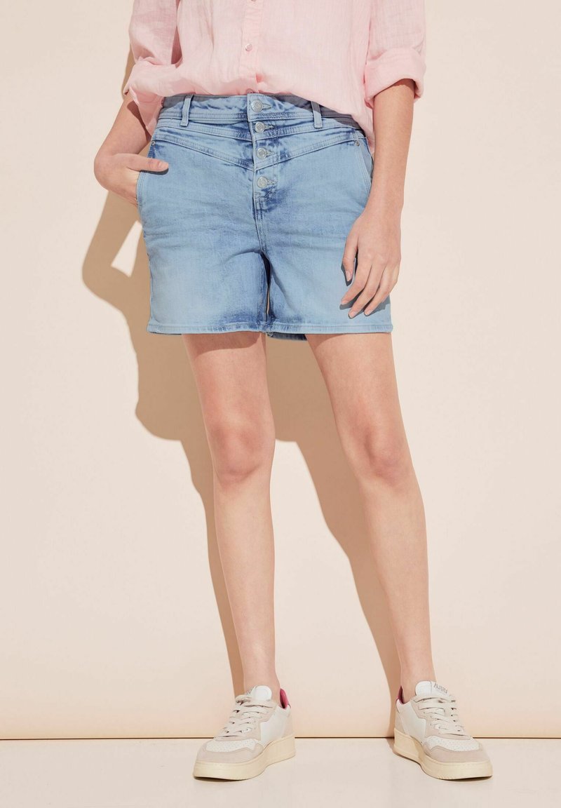 Pantalones cortos de mezclilla azul claro con cierre de cinco botones, bolsillos laterales y un ajuste casual, combinados con zapatillas blancas con acentos rosas.