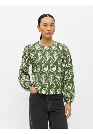 Femme aux cheveux foncés attachés, portant un chemisier vert avec une broderie florale blanche et un pantalon noir, debout avec une main dans la poche.
