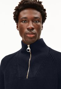 Junger Mann mit kurzem lockigem Haar, der einen navy-blauen gerippten Pullover mit hohem Kragen und einem silbernen Reißverschluss trägt.