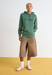 Hanorac verde cu logo purpuriu, buzunar frontal; pantaloni scurți largi maro; adidași roz cu accente albe și purpurii; fundal simplu.